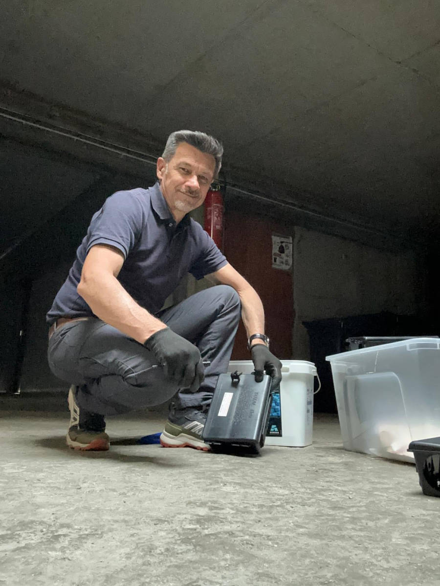 Technicien ACS38 accroupi dans un sous-sol, gants aux mains, installant un poste d'appâtage sécurisé